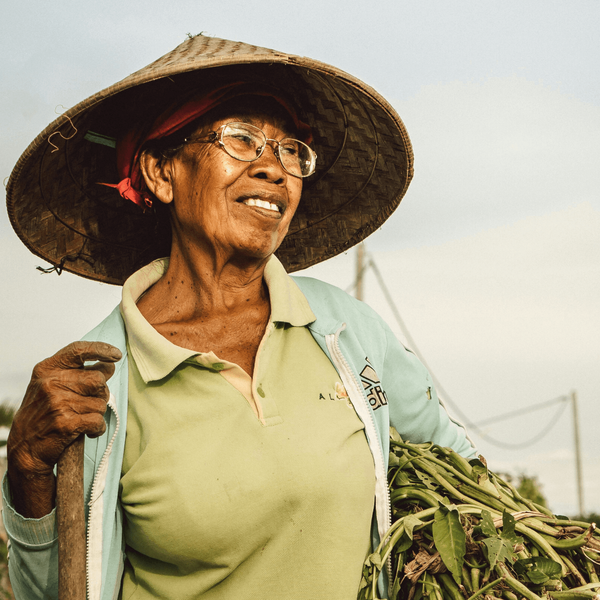 Voyage en Asie — Portrait d'une femme indonésienne âgée portant un chapeau conique traditionnel, souriante et en plein travail agricole à Bali, dans un cadre rural authentique.
