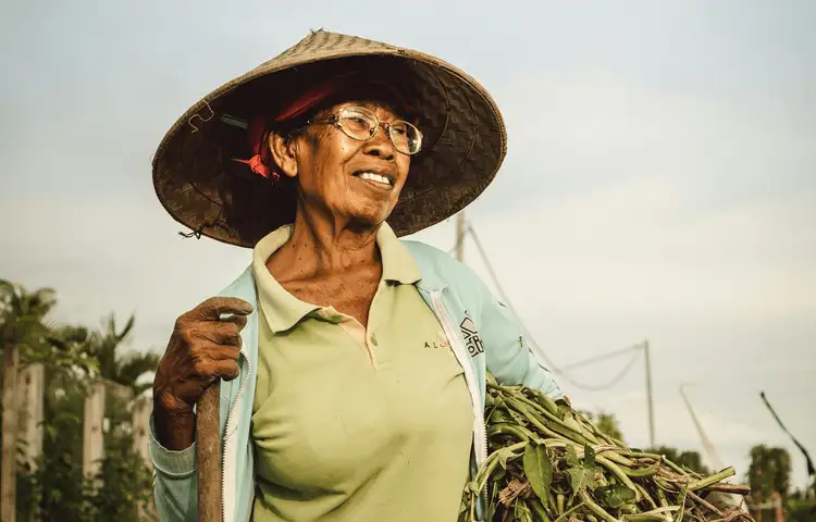 Voyage en Asie — Portrait d'une femme indonésienne âgée portant un chapeau conique traditionnel, souriante et en plein travail agricole à Bali, dans un cadre rural authentique.