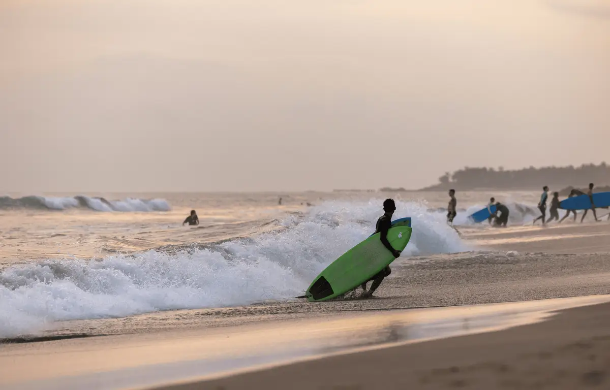 Voyage en Asie — Surfeurs à Bali au coucher du soleil sur une plage avec vagues et sable foncé.