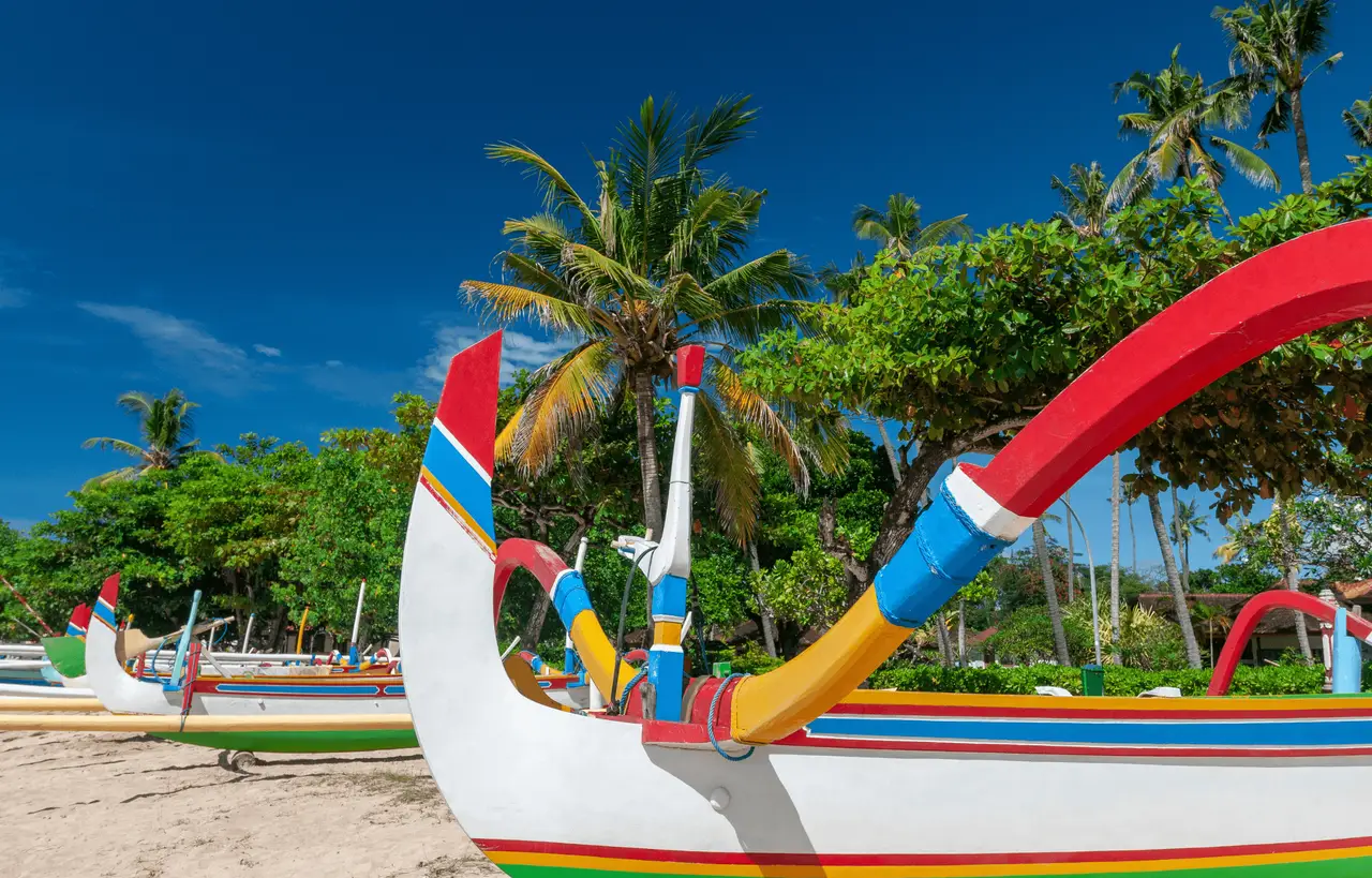 Plusieurs bateaux de pêche traditionnels balinais, appelés jukung, alignés sur le sable d'une plage bordée de cocotiers sous un ciel bleu éclatant à Sanur.