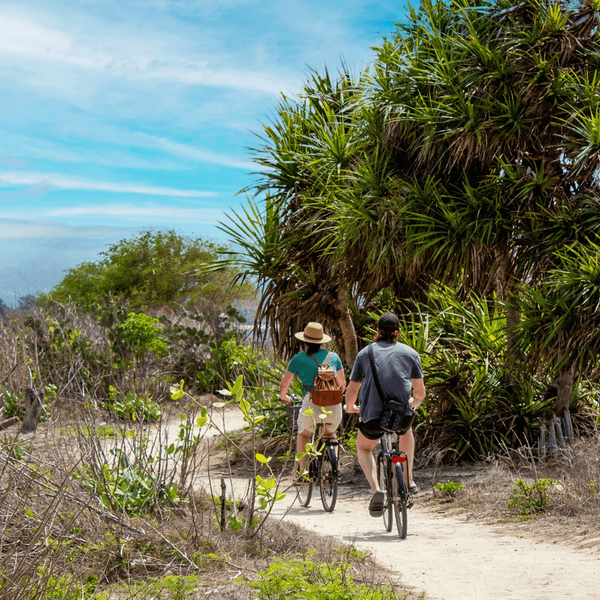 Voyage en Asie — Cycliste balinais en sarong et caping sur un vélo dans un village avec maisons en bambou et chaume.
