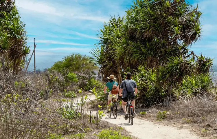 Voyage en Asie — Cycliste balinais en sarong et caping sur un vélo dans un village avec maisons en bambou et chaume.