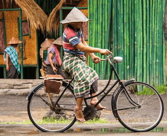 Voyage en Asie — Cycliste balinais portant un sarong traditionnel et un chapeau conique (caping) dans un village, circulant sur un vélo de ville avec sacoche, illustrant la vie locale et rurale.