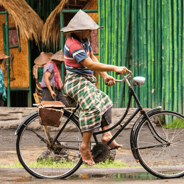 Voyage en Asie — Cycliste balinais portant un sarong traditionnel et un chapeau conique (caping) dans un village, circulant sur un vélo de ville avec sacoche, illustrant la vie locale et rurale.