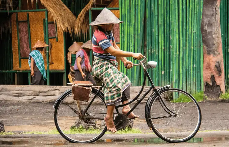 Voyage en Asie — Cycliste balinais portant un sarong traditionnel et un chapeau conique (caping) dans un village, circulant sur un vélo de ville avec sacoche, illustrant la vie locale et rurale.