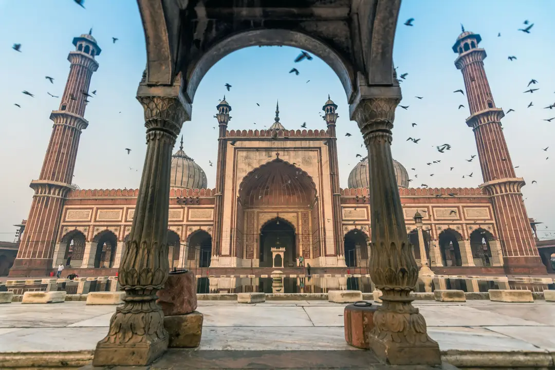 La majestueuse mosquée Jama Masjid en grès rouge et marbre blanc à Old Delhi, photographiée à travers une arche en pierre sculptée alors que des oiseaux s'envolent dans le ciel.