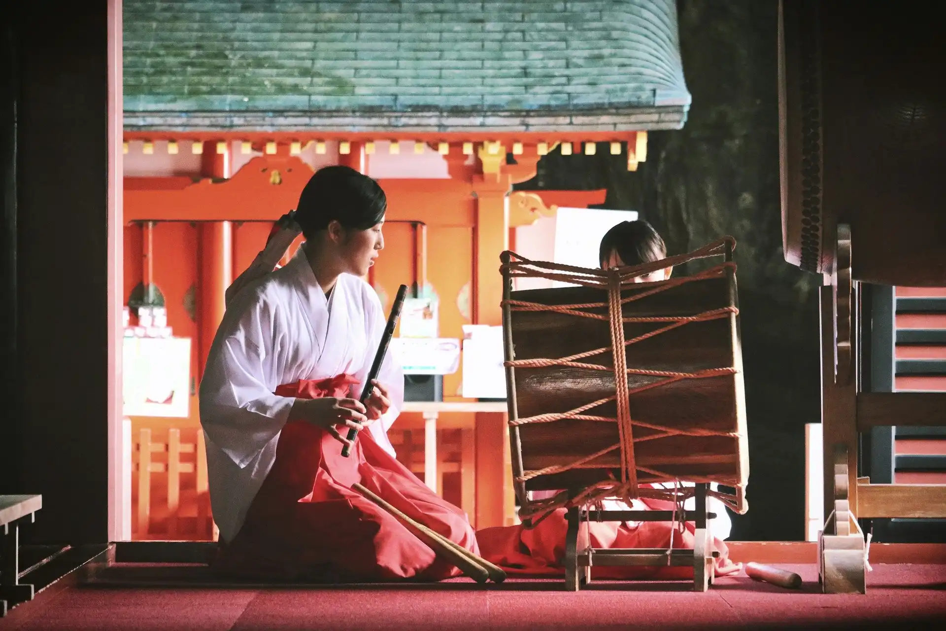 traditional japanese musician in white robe playing a flute inside the sacred red miyazaki jingu shrine surrounded by ancient forest