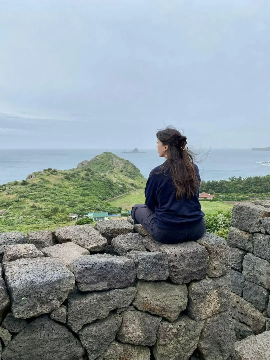 Une touriste de dos, assise sur un muret de pierres volcaniques, contemple les collines verdoyantes et le panorama marin de la côte de Yongmeori à Jeju.