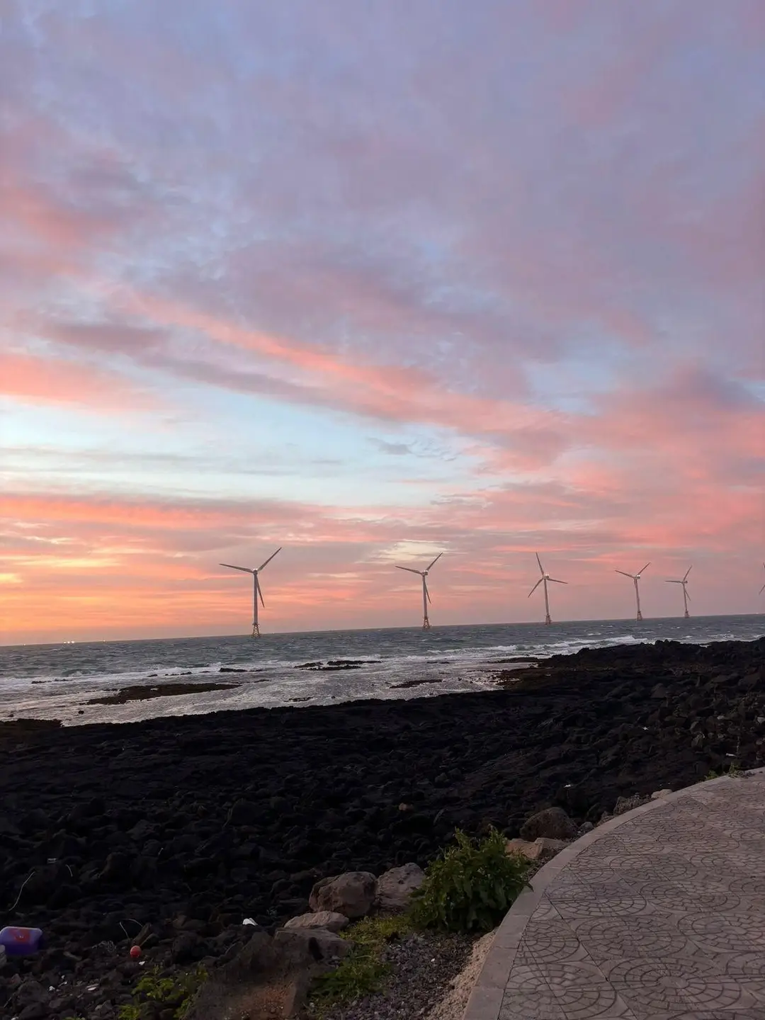 Coucher de soleil aux nuances de rose et d'orange sur une côte de roches volcaniques noires, avec une rangée d'éoliennes offshore à Jeju.