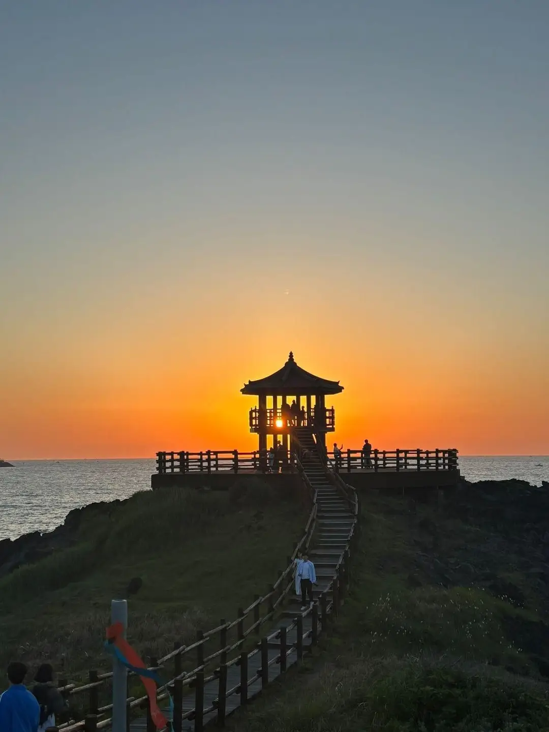 Silhouette d'une pagode traditionnelle en bois construite sur une avancée rocheuse face à la mer, traversée par les lueurs d'un coucher de soleil orange à Jeju.