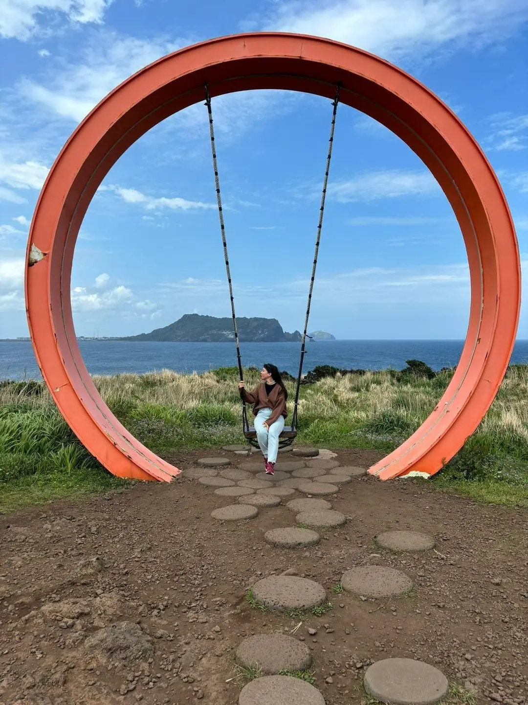 Une touriste assise sur une grande balançoire installée dans une structure circulaire orange, offrant un panorama sur la mer et une île au loin à Jeju.