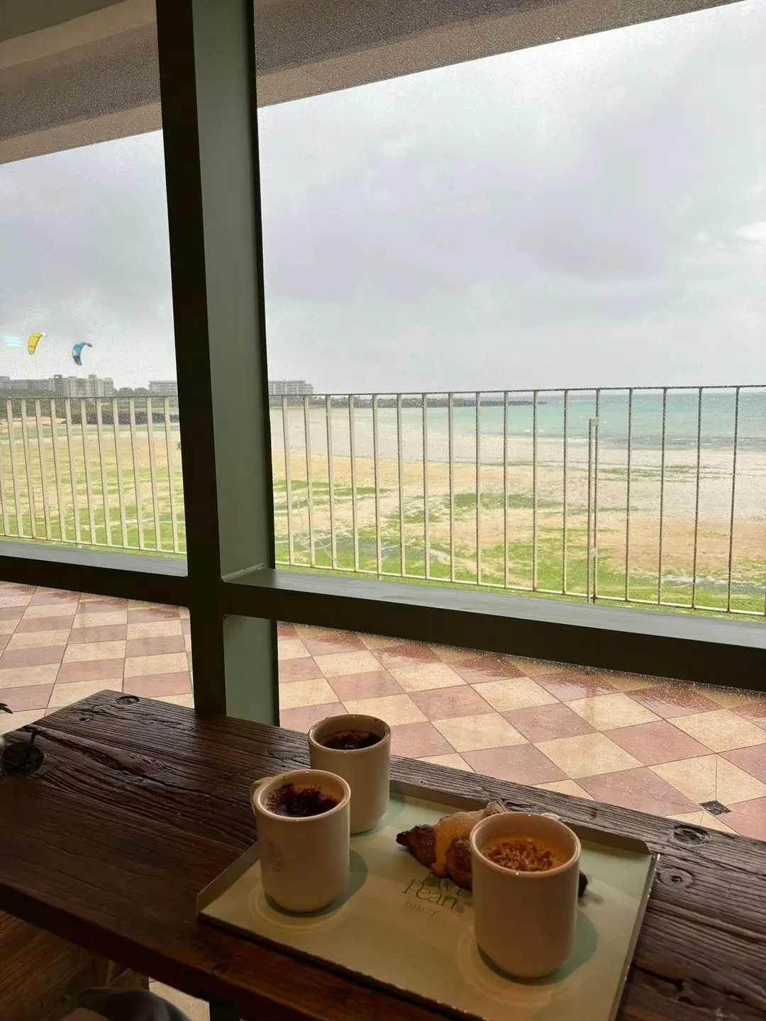 Un plateau de boissons chaudes et viennoiseries posé sur une table en bois face à une grande baie vitrée, offrant une vue sur la plage et des kitesurfs à Jeju.
