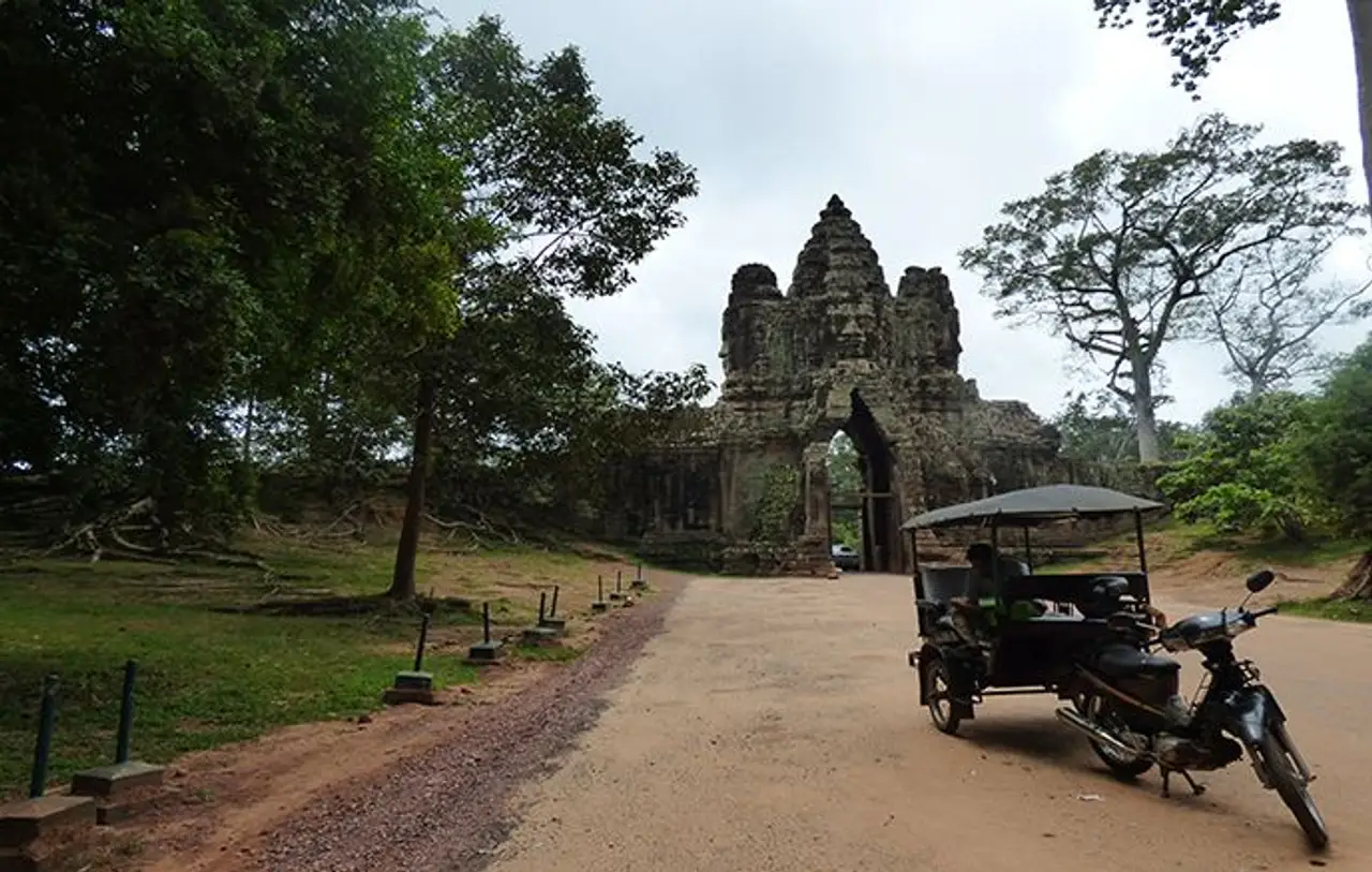 Un tuk-tuk traditionnel garé sur un sentier en terre devant la monumentale porte d'entrée en pierre sculptée de la cité d'Angkor Thom au Cambodge.