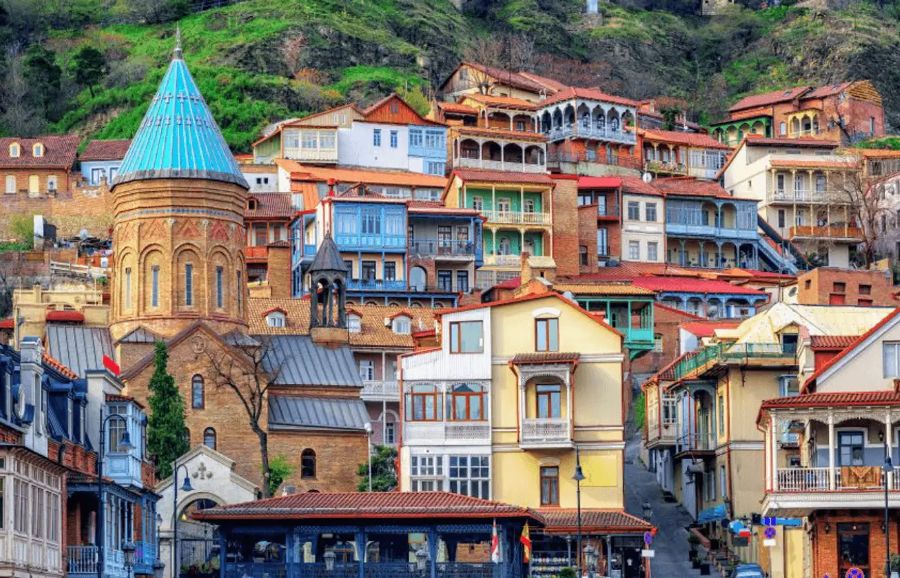 Façades en bois sculpté et balcons colorés des maisons traditionnelles de la vieille ville de Tbilissi en Géorgie, dominées par le dôme conique bleu d'une église orthodoxe à flanc de colline.