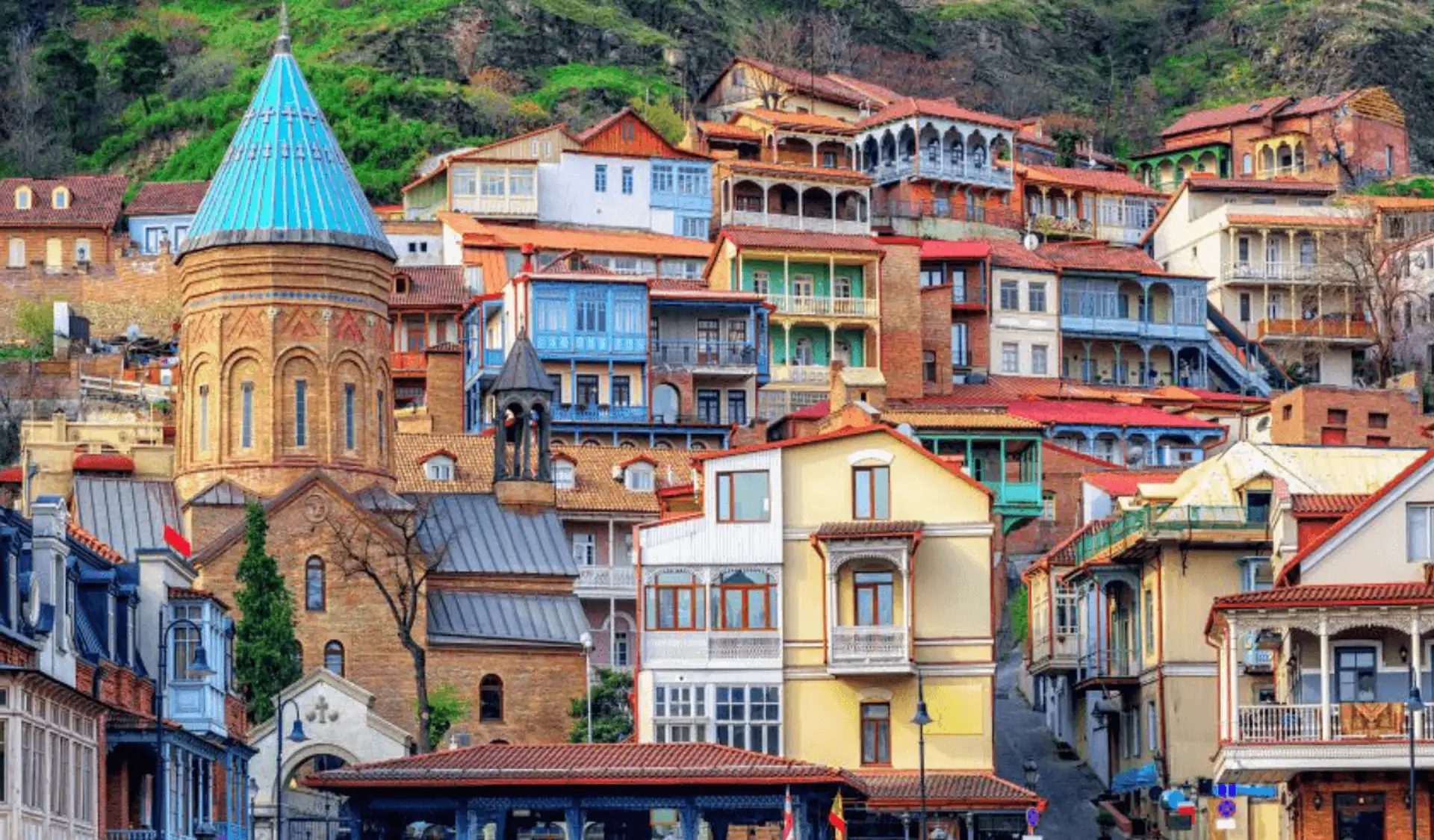 Façades en bois sculpté et balcons colorés des maisons traditionnelles de la vieille ville de Tbilissi en Géorgie, dominées par le dôme conique bleu d'une église orthodoxe à flanc de colline.