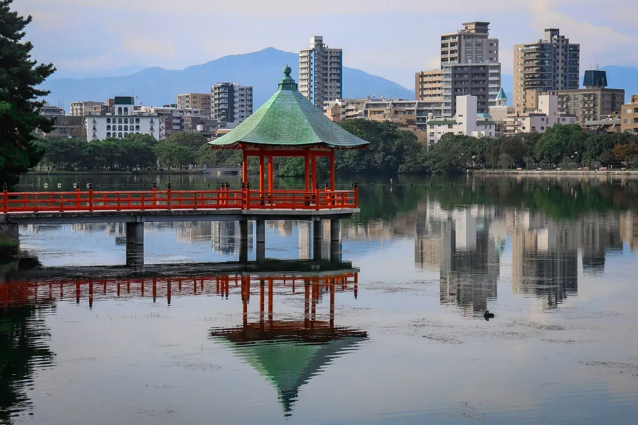 traditional hexagonal wooden pavilion on a lake with a red bridge overlooking modern buildings in ohori park fukuoka