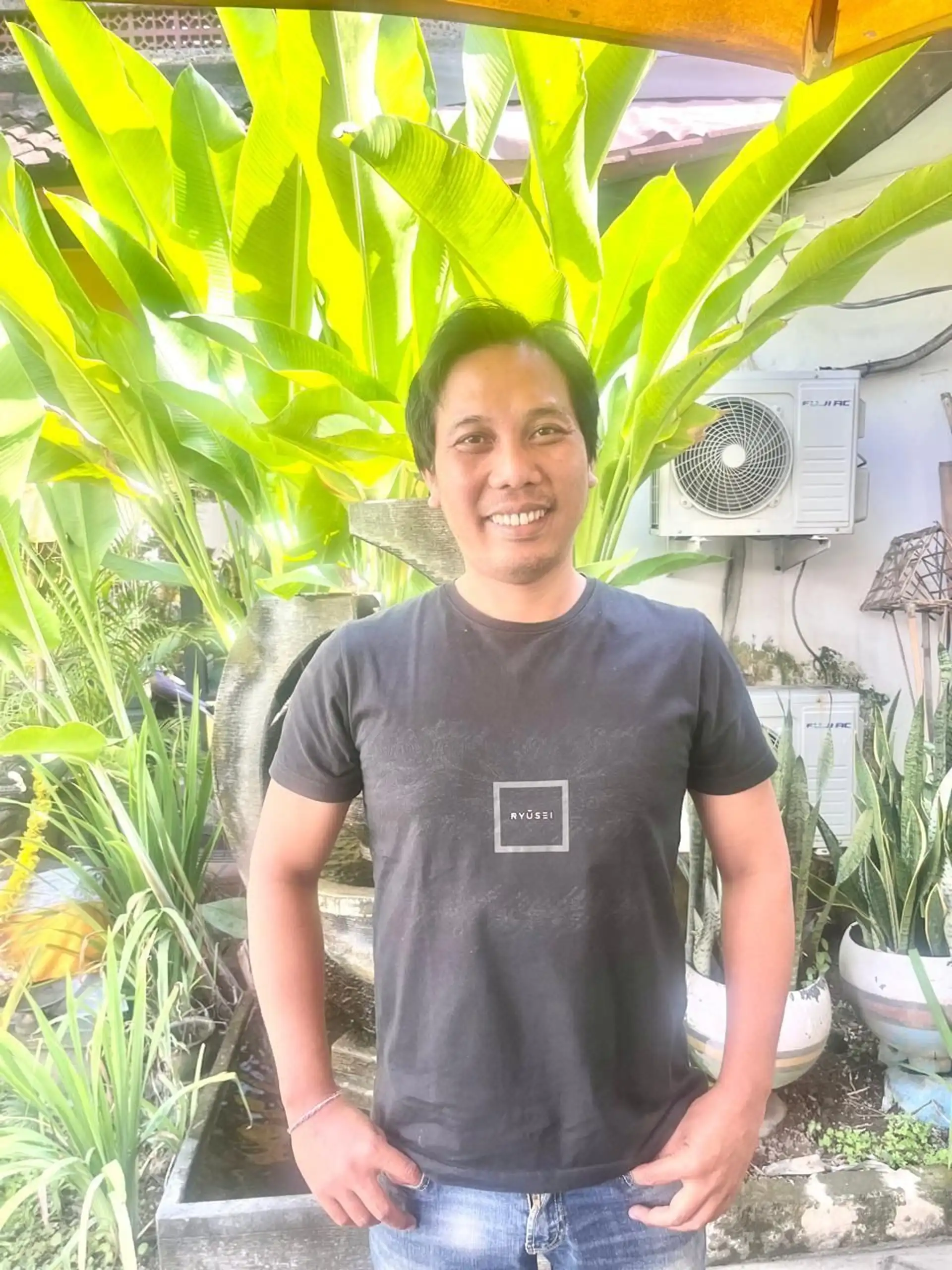 Portrait de Komang, responsable des opérations sur le terrain pour Shanti Travel en Indonésie, souriant devant des plantes tropicales à Bali.