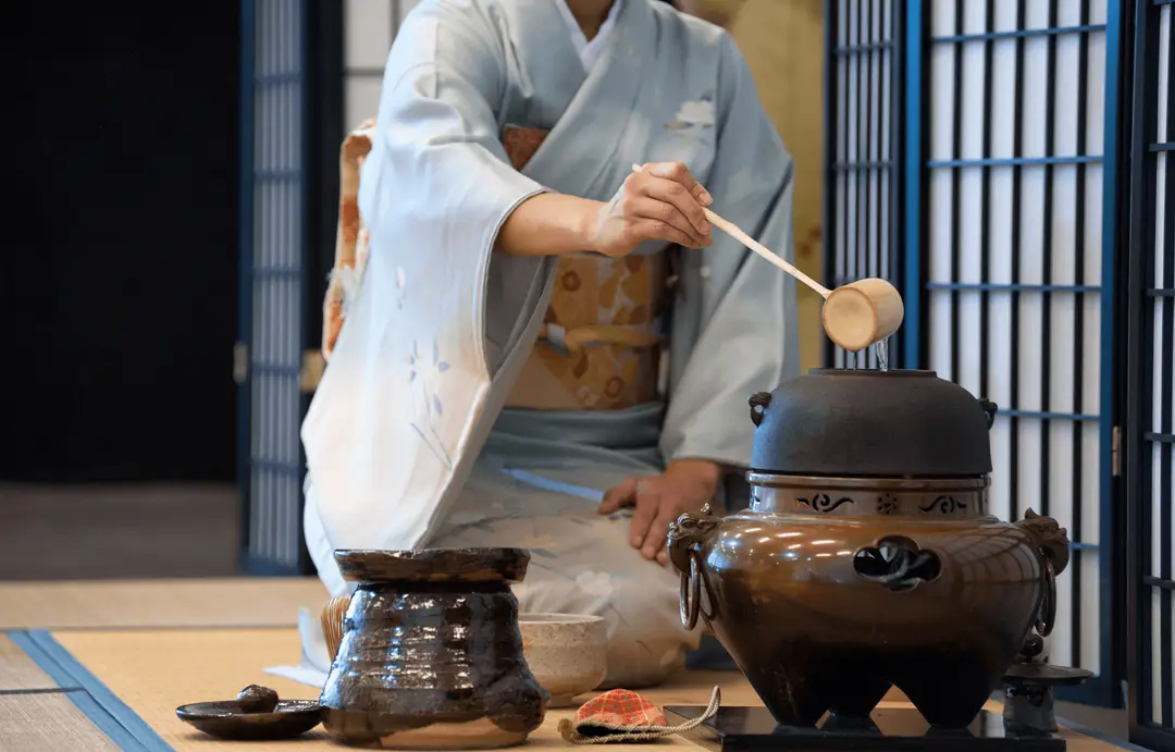 Route du thé au Japon – Femme en kimono bleu pastel puisant de l'eau chaude dans un kettle en fonte avec une louche en bambou.