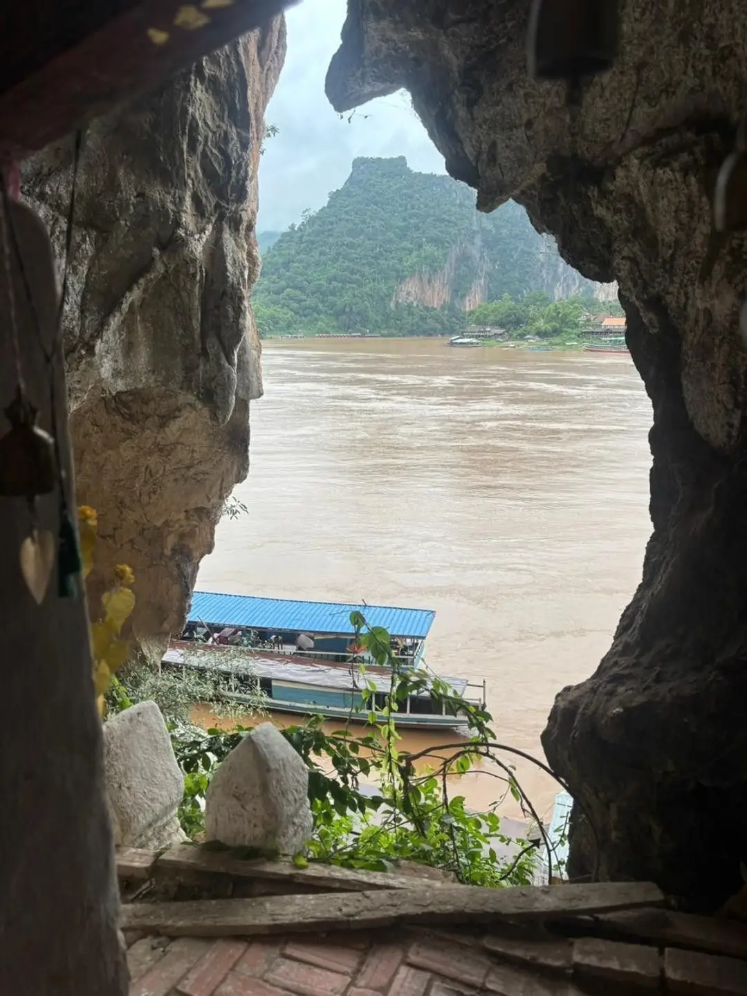 Vue depuis l'ouverture rocheuse de la grotte de Pak Ou donnant sur le fleuve Mékong, avec un bateau de croisière amarré en contrebas et des falaises verdoyantes au loin.
