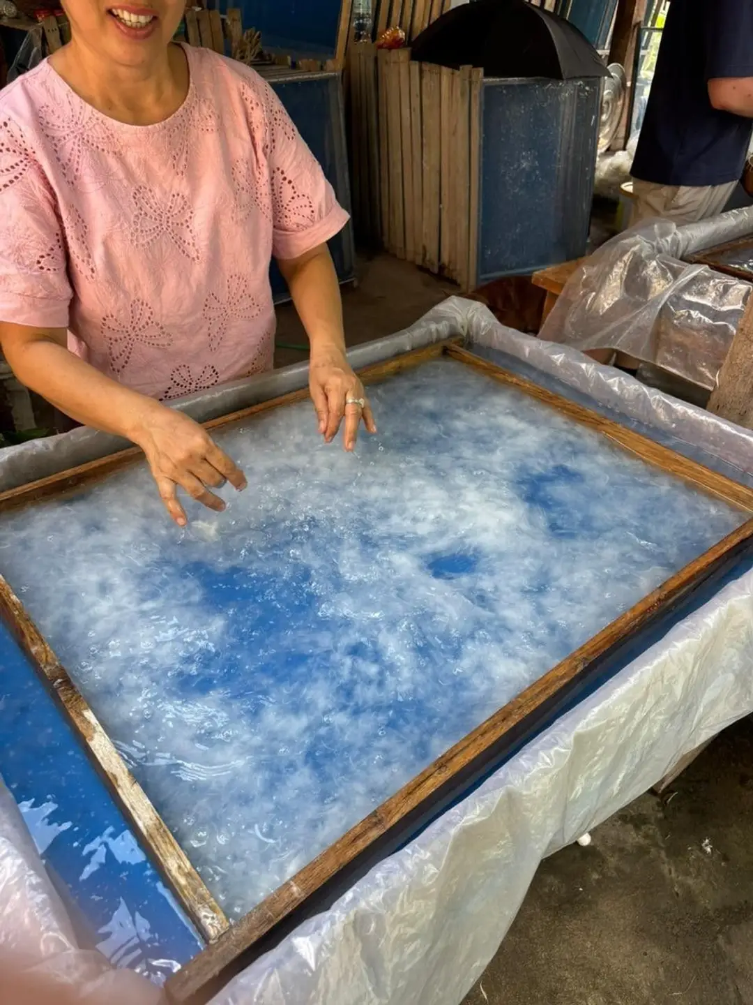 Une artisane montre le processus de fabrication du papier traditionnel dans un grand bac d'eau bleue où flottent les fibres de cellulose blanches réparties sur un tamis en bois.