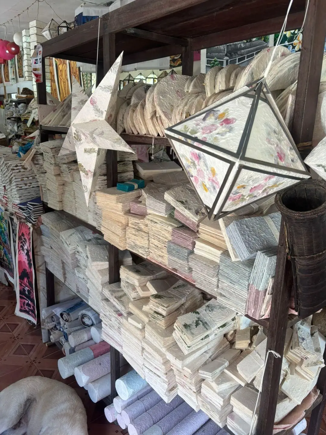 Des étagères en bois remplies de carnets artisanaux, de feuilles empilées et de lanternes décoratives en papier de mûrier fait main avec des motifs floraux.