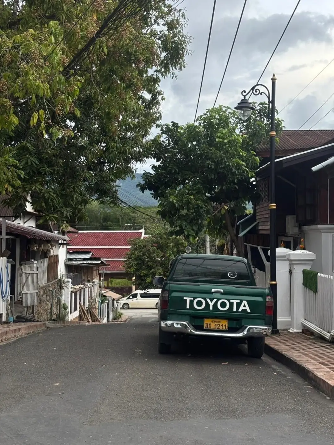 Un pick-up vert garé le long d'une rue paisible et goudronnée bordée de maisons traditionnelles et de verdure dans le centre historique de Luang Prabang.