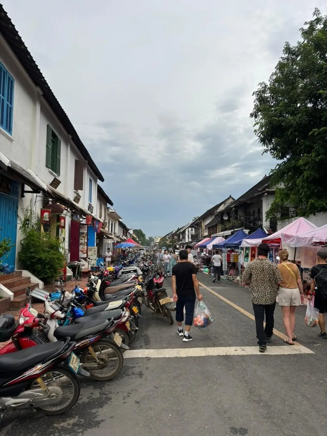 Des voyageurs et des locaux se promènent dans une rue animée bordée de nombreux scooters garés et de stands de marché sous des tentes colorées à Luang Prabang.