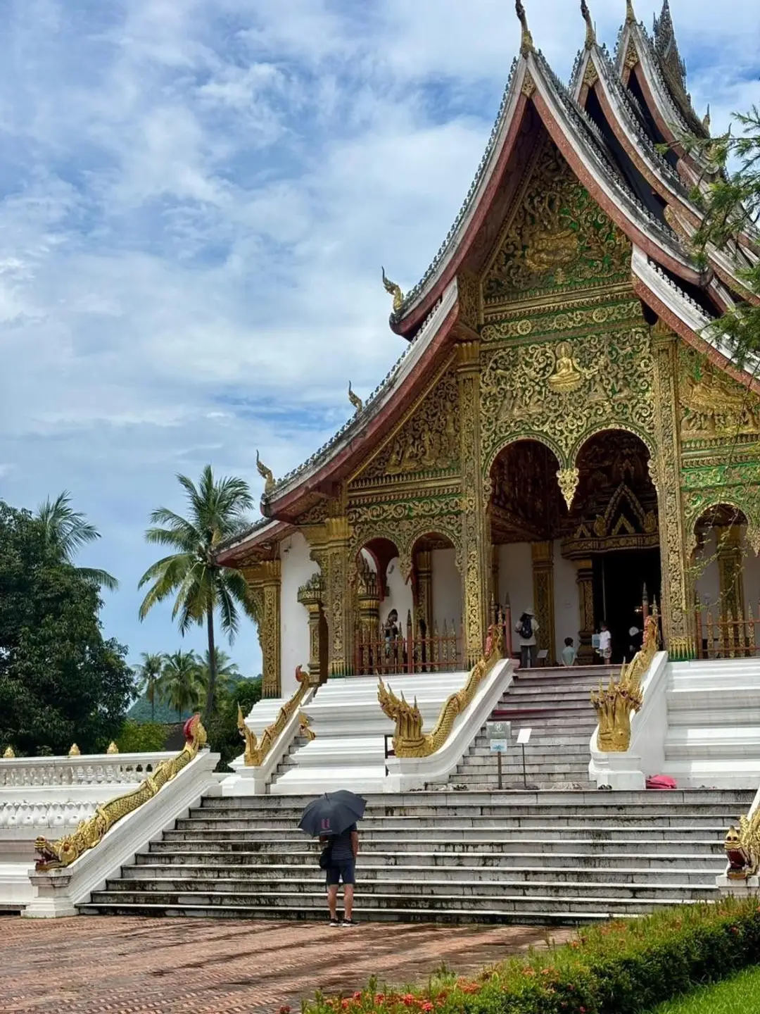 Un visiteur avec un parapluie noir se tient devant le grand escalier blanc menant à la façade richement ornée d'or et de sculptures du temple Haw Pha Bang à Luang Prabang.
