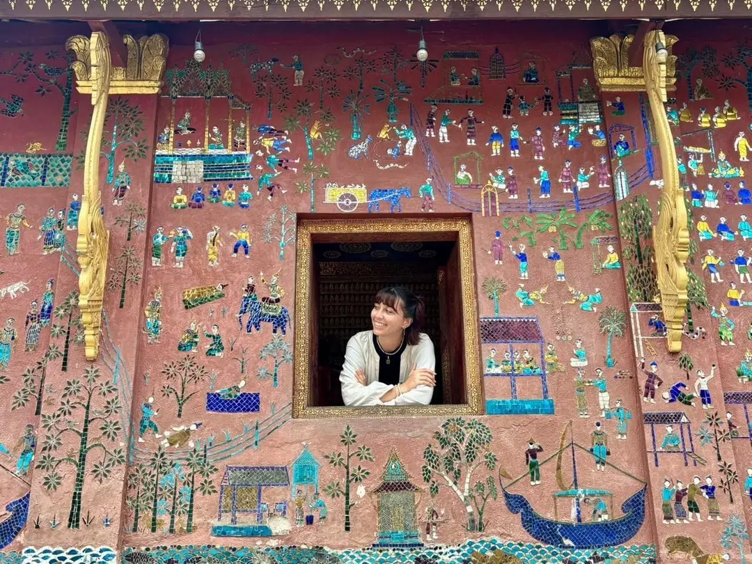 Une voyageuse sourit à une fenêtre intégrée dans un grand mur rouge orné de riches mosaïques colorées représentant des scènes de vie traditionnelles dans un temple de Luang Prabang.