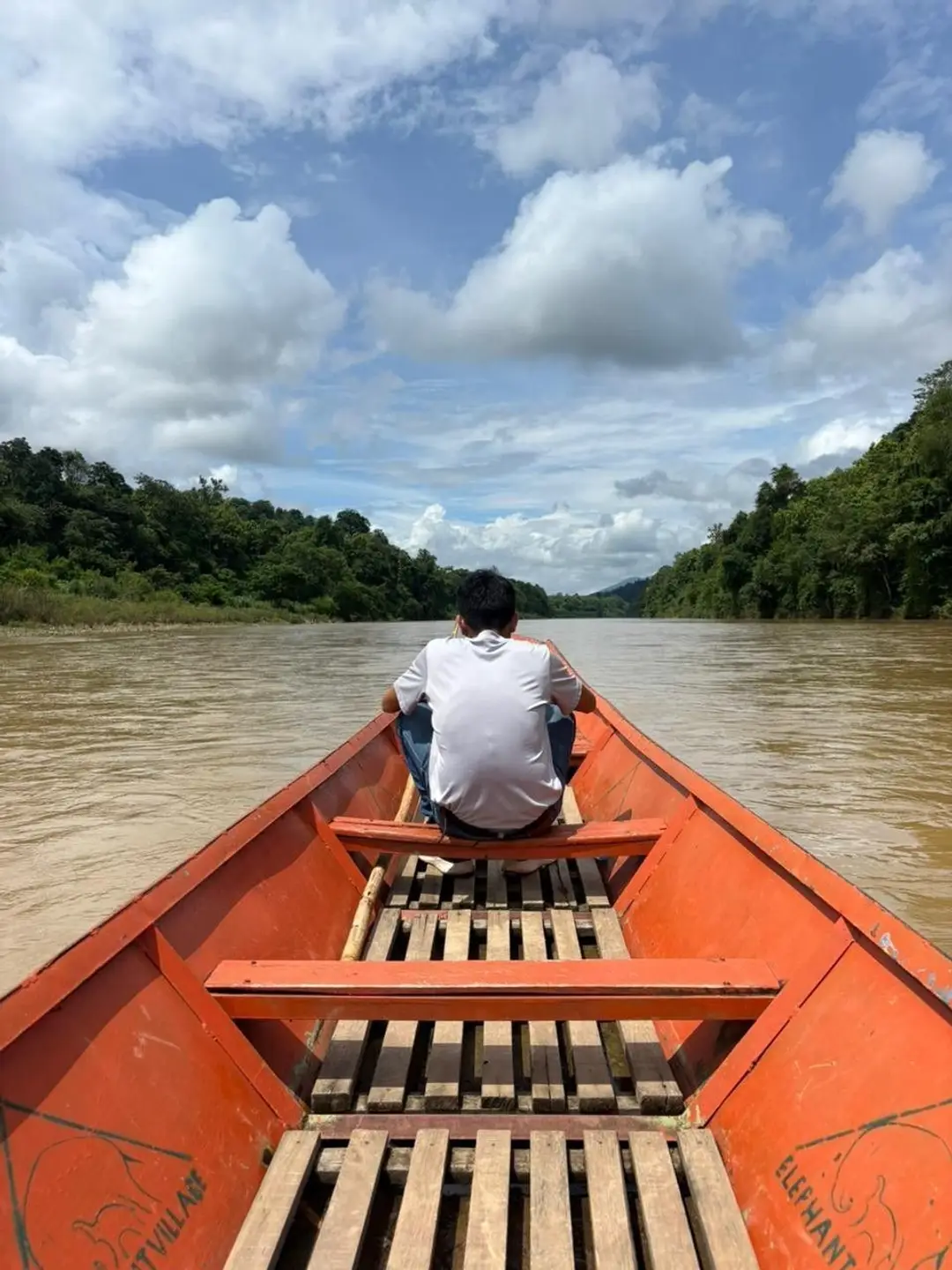 Vue depuis l'intérieur d'une pirogue traditionnelle orange où un passager est assis à l'avant, naviguant sur les eaux calmes et brunes du Mékong bordé de forêts tropicales au Laos.