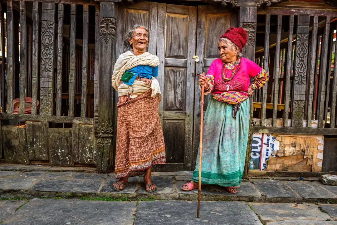 Deux femmes népalaises en tenue traditionnelle souriant devant l'entrée en bois sculpté d'un hébergement local au Népal.