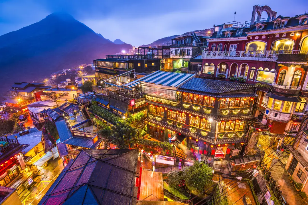 Vue panoramique nocturne du village de montagne de Jiufen à Taïwan, avec ses bâtiments traditionnels illuminés de lumières chaudes et dorées face à la mer sous un ciel de crépuscule.