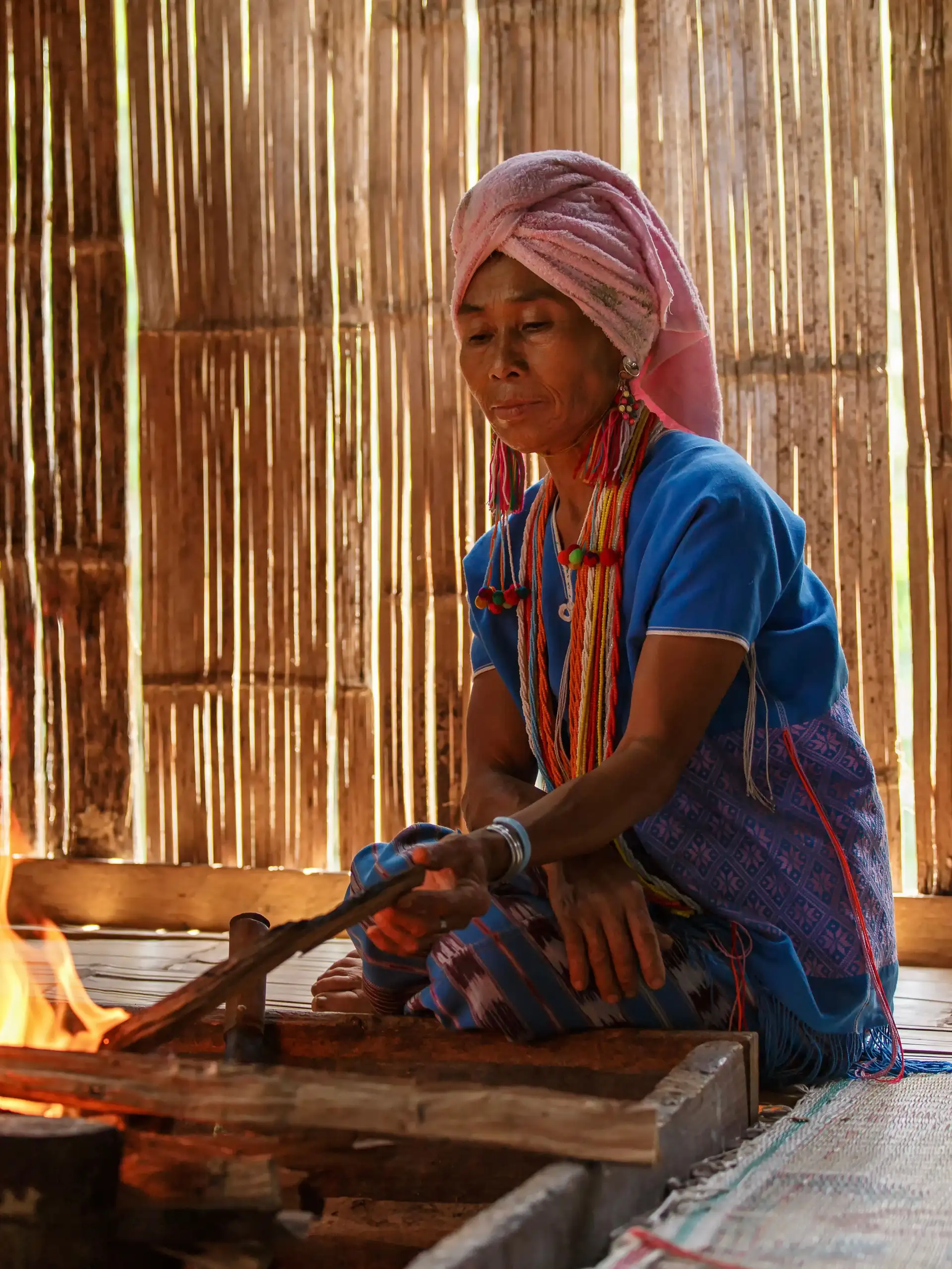 Portrait d'une femme de la tribu Karen en tenue traditionnelle bleue et coiffe rose, portant des colliers en spirale en laiton autour du cou dans un atelier en bambou.