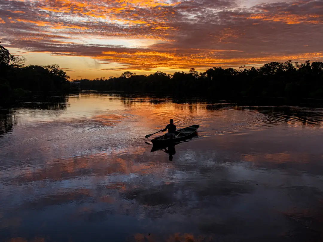 Un pêcheur rame sur un bateau à travers le fleuve Amazone au coucher du soleil, entouré de reflets vifs