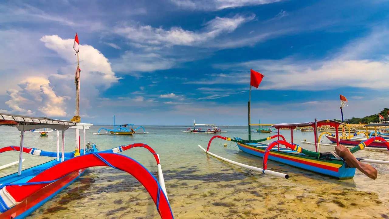 Voyage en Asie – Des jukung, bateaux de pêche traditionnels balinais, alignés sur la plage sous un ciel bleu parsemé de nuages à Bali en Indonésie.