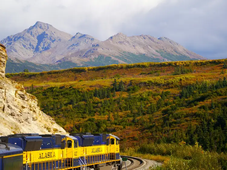 Train jaune et bleu circulant au pied d'une falaise avec vue sur les collines de toundra et de hautes montagnes en Alaska