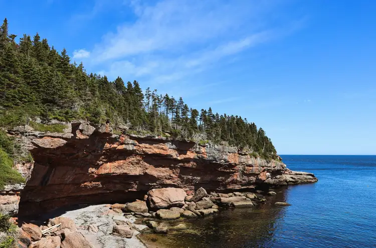 Falaises de roche rougeâtre recouvertes d'une forêt de conifères plongeant dans les eaux bleues de la mer à Percé