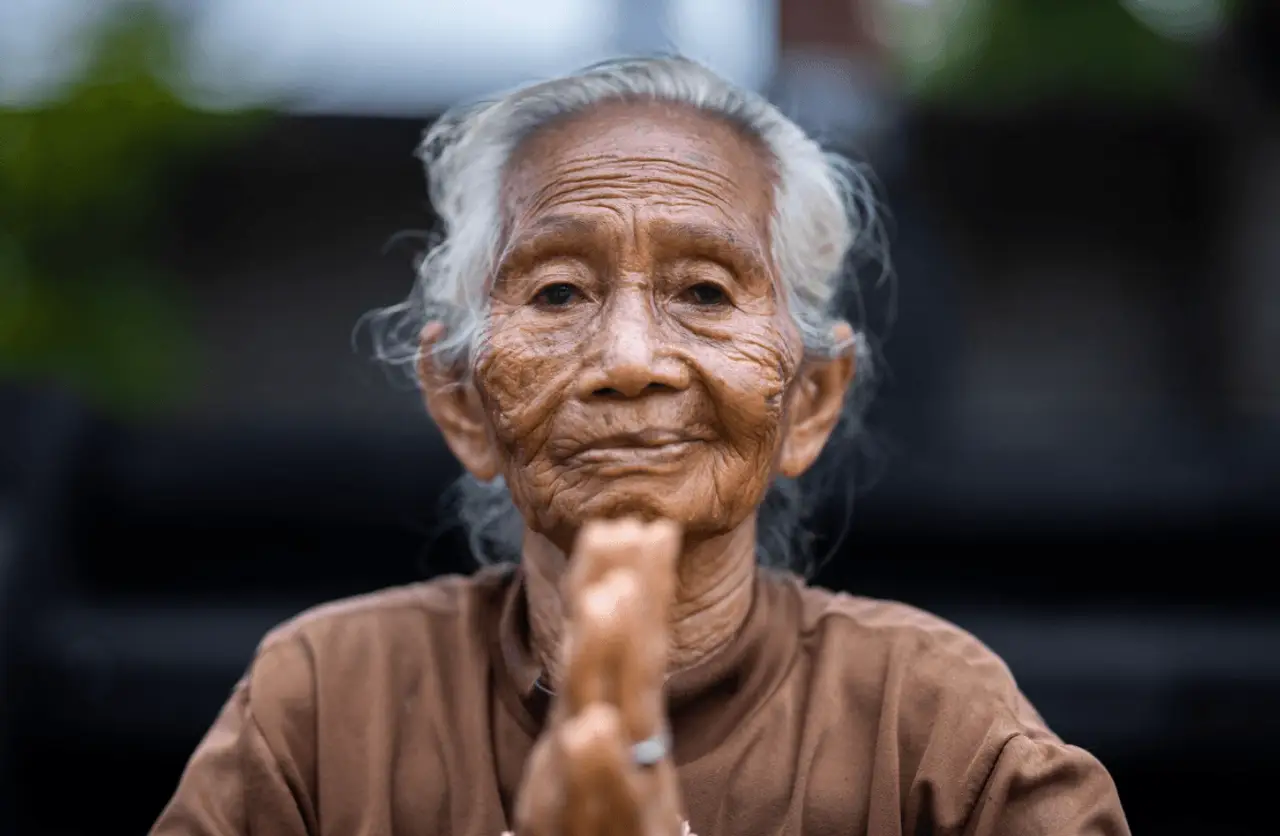 Portrait en gros plan d'une femme âgée balinaise aux cheveux blancs, souriant avec bienveillance et joignant les mains en signe de salut traditionnel pour accueillir les voyageurs à Bali.