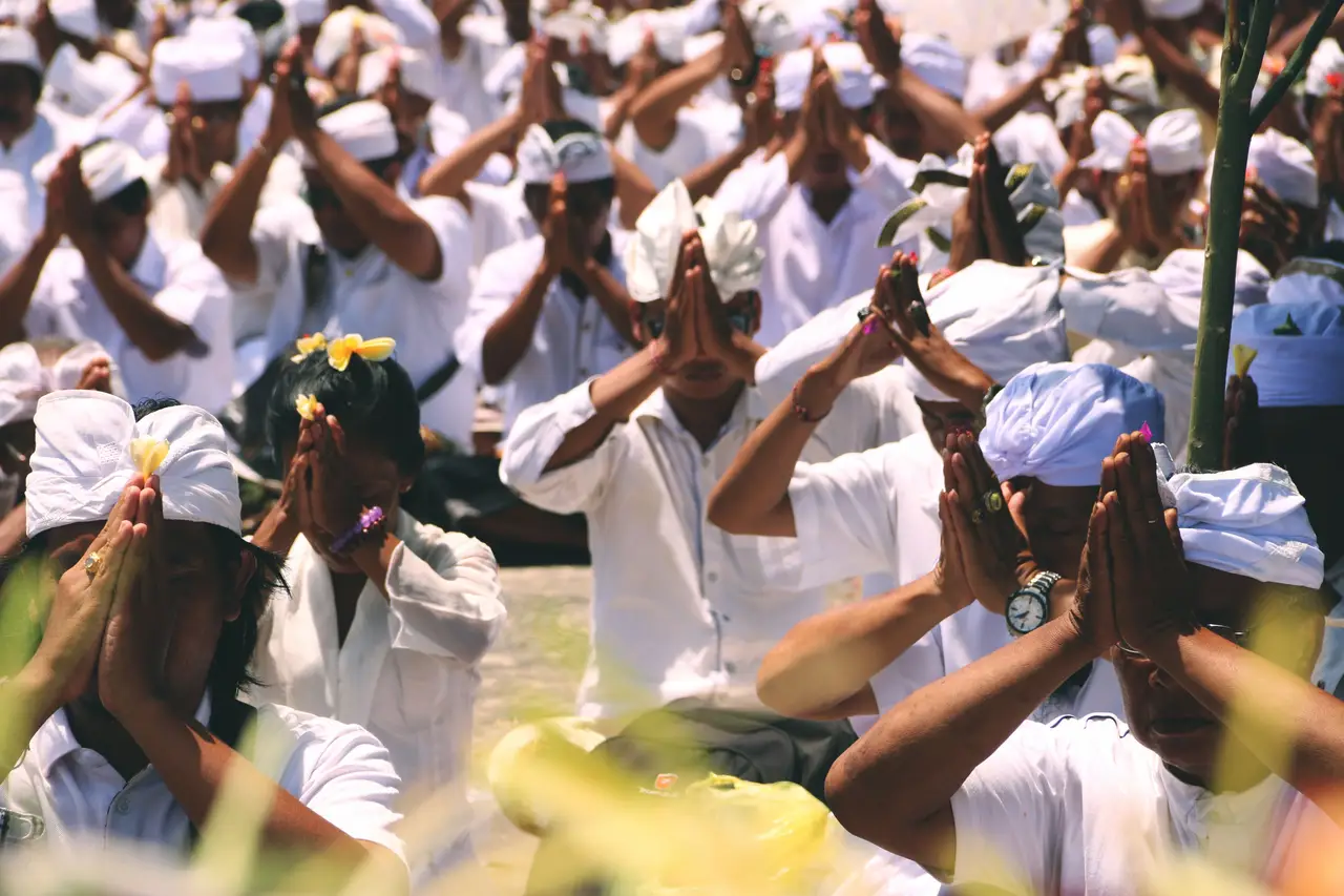 Une foule de fidèles balinais vêtus de blanc et de coiffes traditionnelles udeng, réunis les mains jointes en position de prière lors d'une cérémonie culturelle à Bali.