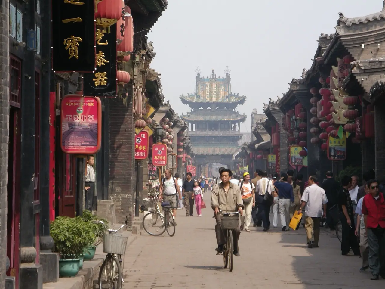 Une perspective sur une ruelle pavée et animée de la vieille ville de Pingyao, bordée de maisons traditionnelles en bois avec des lanternes rouges et une tour fortifiée au loin.