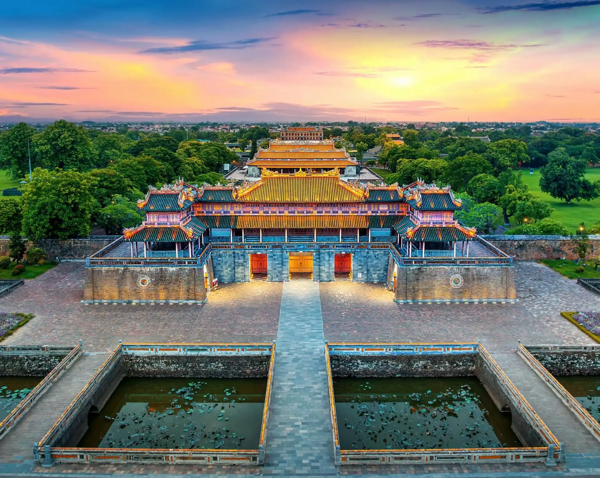 Voyage au Vietnam – Vue panoramique de la porte fortifiée de la Cité Impériale de Hué entourée de verdure et de douves sous un ciel de coucher de soleil.