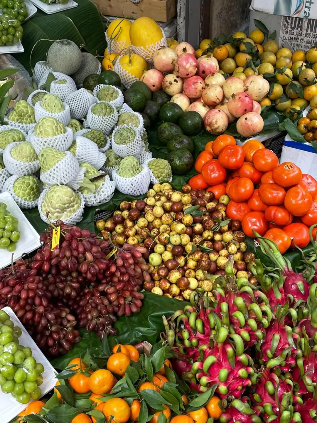 Un étal coloré de marché local présentant une grande variété de fruits frais, dont des fruits du dragon, des pommes cannelle emballées, des raisins et des agrumes.