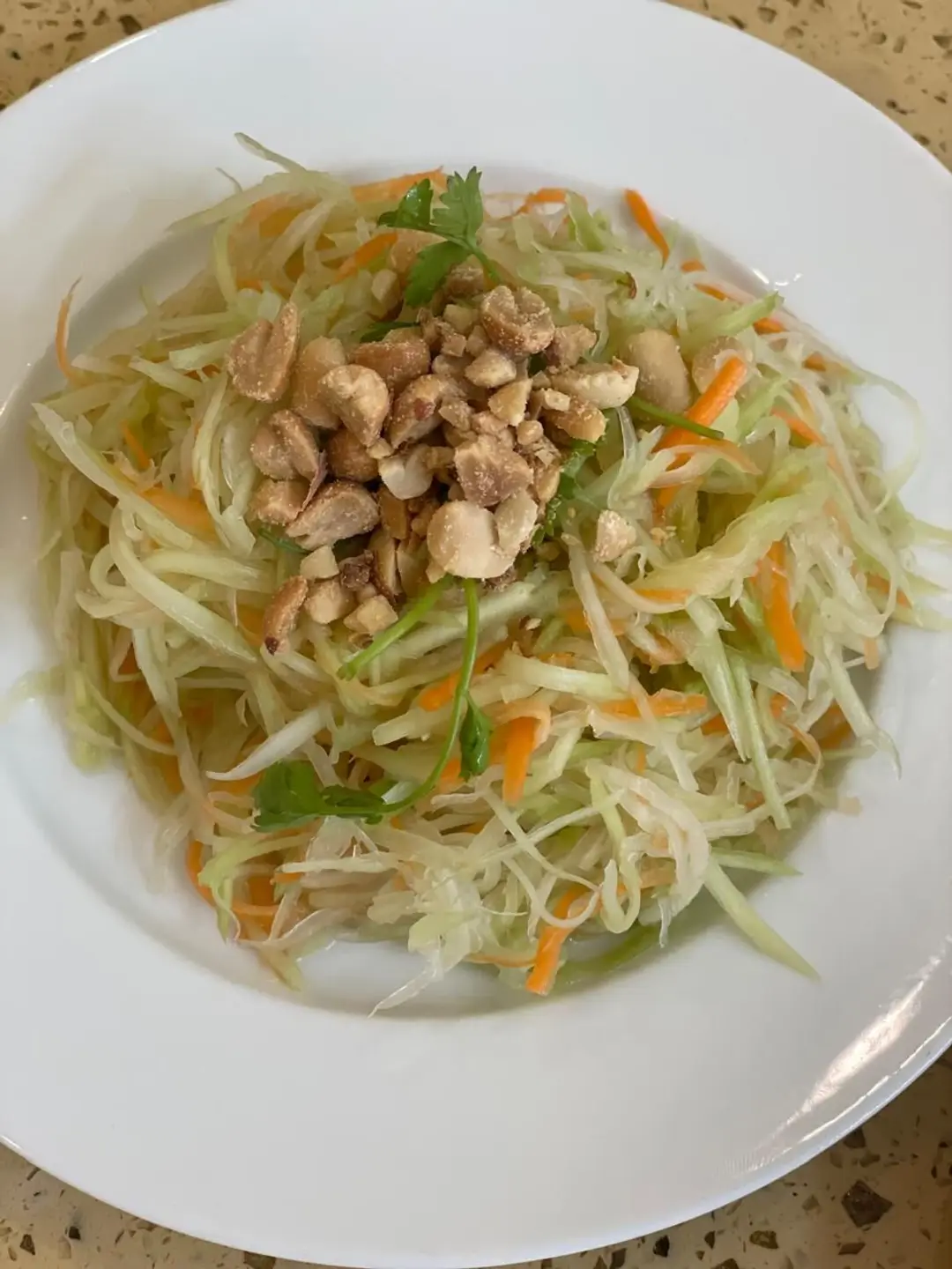 Une assiette blanche contenant une salade traditionnelle de papaye verte râpée et de carottes, parsemée de cacahuètes concassées et d'herbes fraîches au Vietnam.