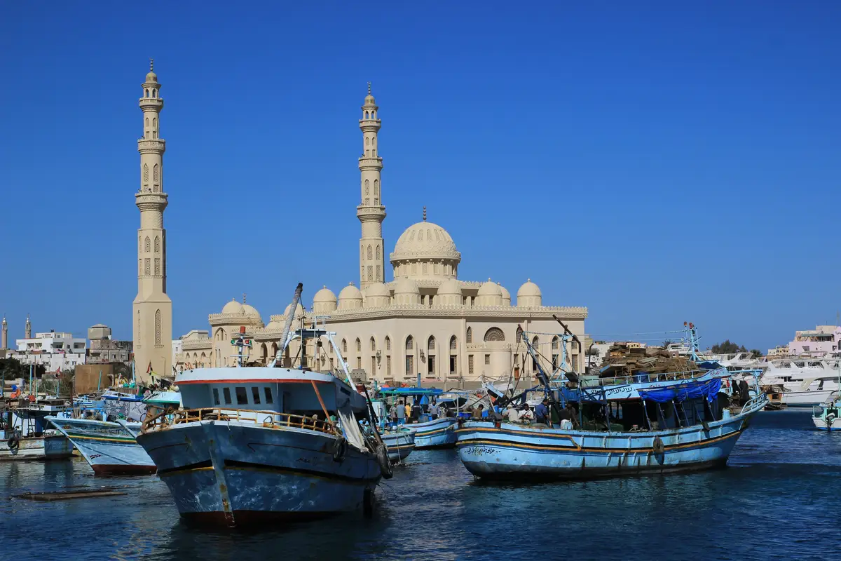 Plusieurs bateaux de pêche traditionnels amarrés dans le port d'Hurghada, avec une grande mosquée et ses minarets sculptés en arrière-plan sous un ciel bleu intense