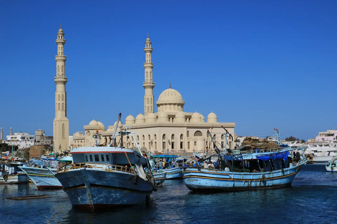 Plusieurs bateaux de pêche traditionnels amarrés dans le port d'Hurghada, avec une grande mosquée et ses minarets sculptés en arrière-plan sous un ciel bleu intense