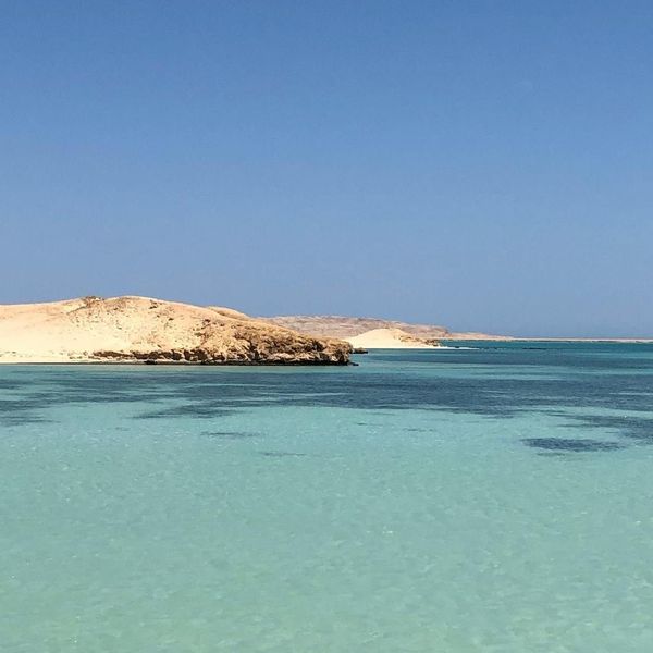 Un panorama paisible d'une plage à Hurghada montrant des eaux turquoise cristallines bordées par des dunes de sable doré sous un ciel bleu sans nuages