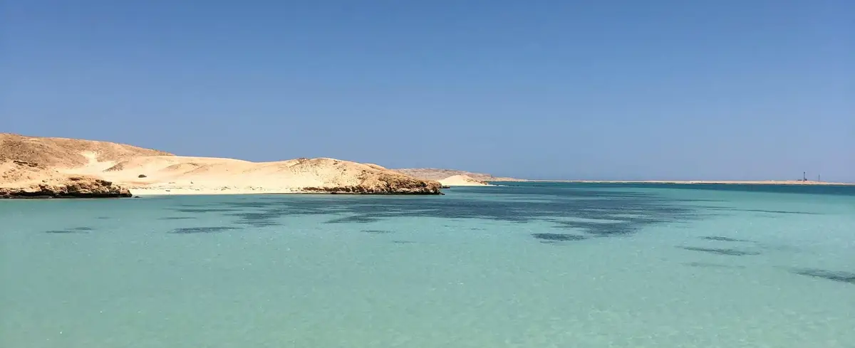 Un panorama paisible d'une plage à Hurghada montrant des eaux turquoise cristallines bordées par des dunes de sable doré sous un ciel bleu sans nuages