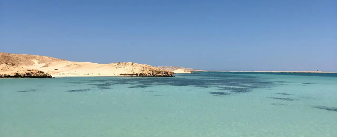 Un panorama paisible d'une plage à Hurghada montrant des eaux turquoise cristallines bordées par des dunes de sable doré sous un ciel bleu sans nuages