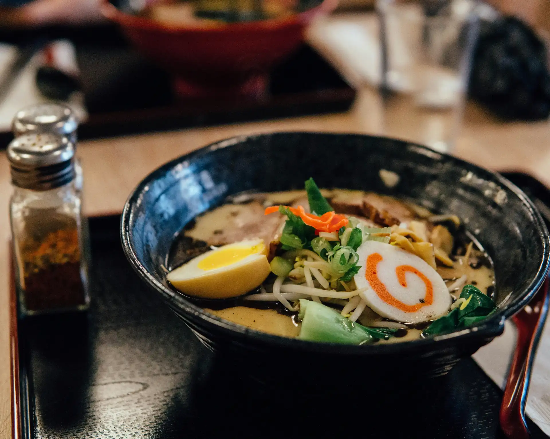 Un bol de ramen traditionnel fumant avec un œuf mollet, du narutomaki et des oignons verts, servi sur une table en bois dans un restaurant au Japon.