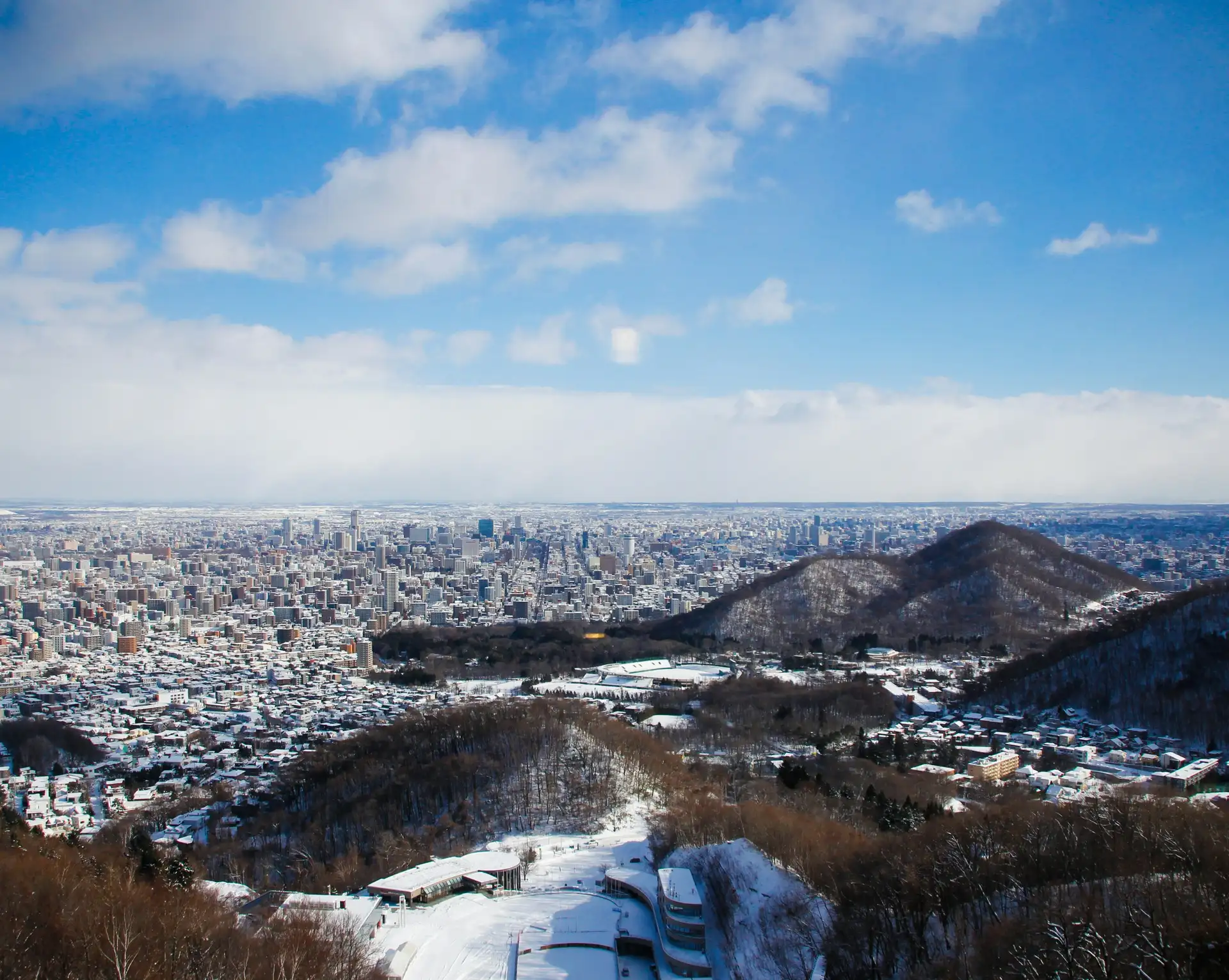 Vue panoramique en plongée sur la ville de Sapporo enneigée depuis une colline, sous un ciel bleu parsemé de nuages blancs à Hokkaido.